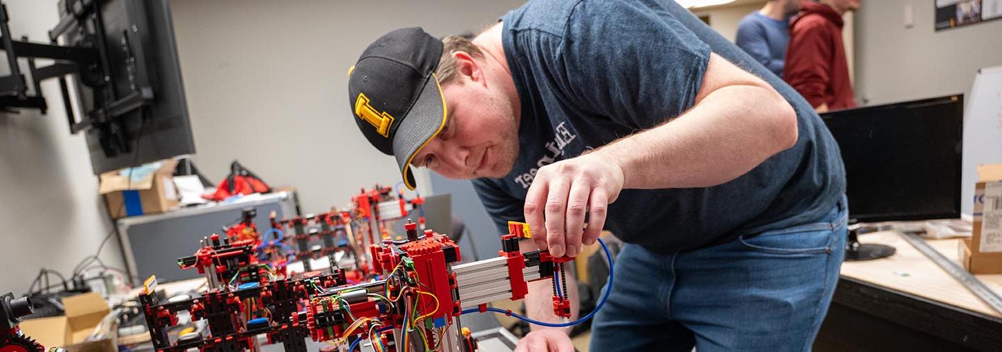 A young man in an Idaho baseball hat adjusts a piece on the arm of a robotic systems that simulates a mini factory in a robotics classroom.